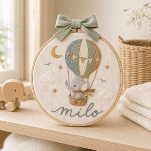 Puede incluir: Un bastidor de bordado circular que presenta un globo aerost&aacute;tico cosido a mano con un elefante gris en la cesta. El dise&ntilde;o incluye una luna creciente amarilla, estrellas, nubes y el nombre "milo" en cursiva, rematado con un lazo de tela verde salvia.