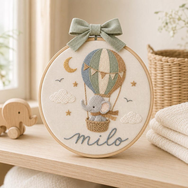 Puede incluir: Un bastidor de bordado circular que presenta un globo aerost&aacute;tico cosido a mano con un elefante gris en la cesta. El dise&ntilde;o incluye una luna creciente amarilla, estrellas, nubes y el nombre "milo" en cursiva, rematado con un lazo de tela verde salvia.