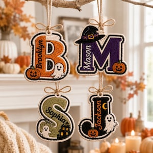 May include: Four hanging ornaments shaped as letters B, M, S, and J. The letter B is orange with the name Brooklyn, a ghost, and a pumpkin. The letter M is purple with the name Mason, a witch hat, and a pumpkin. S is green with Sophia, a ghost, and a house. J is black with Jackson, a ghost, and a pumpkin.