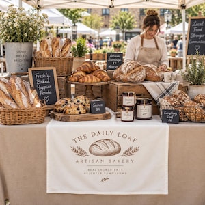May include: A bakery stall display featuring a white table runner with the text The Daily Loaf Artisan Bakery, baskets of baguettes, sourdough loaves, scones, and jars of jam. Small black chalkboards display prices and phrases like Freshly Baked Happier People.