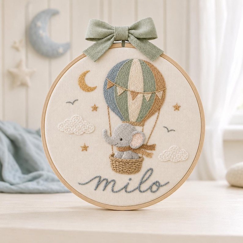 Puede incluir: Un bastidor de bordado circular con un globo aerost&aacute;tico cosido y un elefante gris en la cesta. El dise&ntilde;o incluye una luna creciente amarilla, estrellas, nubes y p&aacute;jaros sobre tela color crema. El nombre milo est&aacute; bordado debajo en azul.