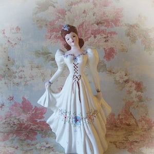 May include: A white porcelain figurine of a woman in a long dress. The dress features a corset-style bodice with brown lacing and is adorned with blue and pink floral accents. The woman has brown hair and is holding the skirt of her dress.