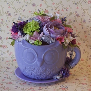 May include: A lavender owl-shaped teacup planter filled with an assortment of colorful artificial flowers. The teacup sits on a matching saucer and features a decorative handle and ribbon. The flowers include purple, pink, green, and white blooms.