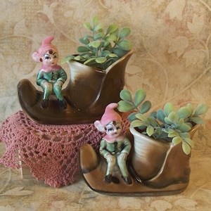Puede incluir: Dos macetas de cerámica con forma de bota de elfo. Cada maceta de bota marrón tiene un interior color crema y contiene una pequeña planta verde. Un elfo de cerámica con sombrero rosa y atuendo verde se sienta en la parte delantera de cada bota.