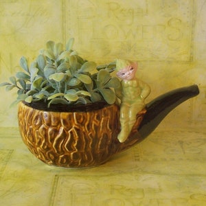 May include: A brown ceramic pipe planter with a green leafy plant and a small green ceramic figurine of a pixie sitting on the pipe.
