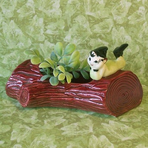 May include: A ceramic planter shaped like a log, painted in a deep red-brown color. The planter holds green faux plants and is adorned with a small figurine of a reclining elf. The elf has a green hat and black shoes.
