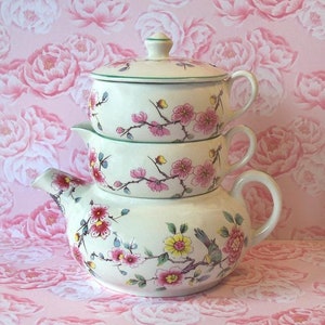 May include: Stacked cream-colored ceramic teapot and serving bowls with pink floral designs and green trim. The teapot has a curved spout and handle. The bowls are stacked on top of each other. The background is a pink floral pattern.