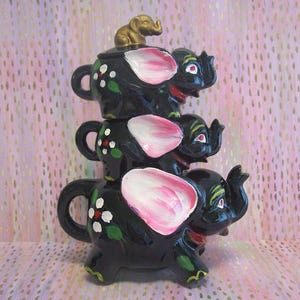 May include: A stack of three black ceramic elephant-shaped teapots with pink ears, green and white floral accents, and red details. The top teapot features a small gold-colored elephant figurine on its lid, and each piece has a handle and spout.