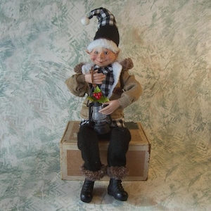 May include: A decorative elf figurine wearing a black and white buffalo plaid hat with a white pom-pom, a tan jacket with white faux fur trim, and black trousers. The elf holds a small black lantern adorned with green leaves and red berries.