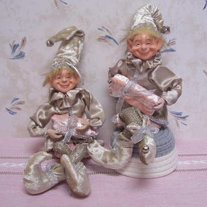May include: Two whimsical elf dolls with smiling faces, wearing gold satin outfits and tall, pointed hats. Each doll holds a pink gift wrapped with silver ribbon. One doll sits on a gray and white woven base.