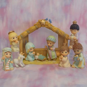 May include: A porcelain nativity set featuring a stable with a gold star, containing figurines of a baby in a manger, Mary, Joseph, an angel, a shepherd with a goat, and three wise figures, all with teardrop-shaped eyes and pastel clothing.