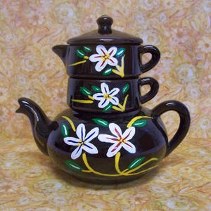 May include: A dark brown ceramic teapot set with a teapot, a lid, and two stacked cups. The set is decorated with white and red flowers with yellow and green accents. The teapot has a curved spout and a rounded handle.