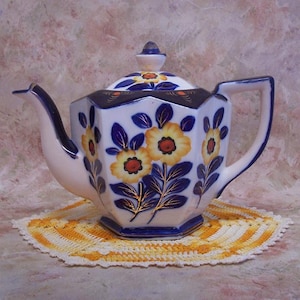 May include: A white ceramic teapot with a hexagonal body, featuring a hand-painted floral pattern of yellow flowers with dark blue leaves and gold accents. The teapot includes a matching lid with a small knob, a curved spout, and a handle.