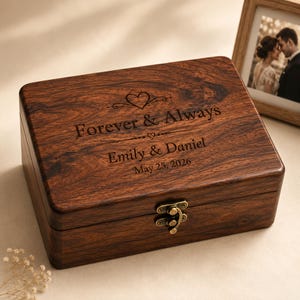 May include: A rectangular dark brown wooden keepsake box with a hinged lid and a brass-coloured metal latch. The lid is engraved with a heart icon, the text "Forever & Always", the names "Emily & Daniel", and the date "May 25, 2026".