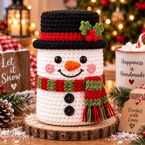 Könnte beinhalten: Eine weiß gehäkelte Schneemannfigur mit einem schwarzen Zylinder mit rotem Band und grünen Stechpalmenblättern, einem rot-grün gestreiften Schal und zwei schwarzen Knöpfen am Körper. Der Schneemann hat schwarze Augen, eine orange Nase und rosa Wangen.