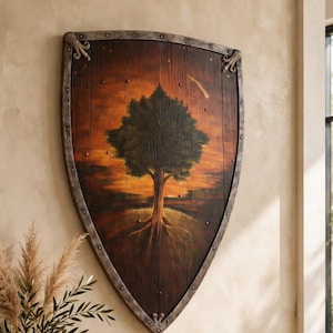 May include: A large, shield-shaped wooden wall hanging featuring a painted scene of a tree against an orange and brown sunset background, framed by a dark metal border with visible rivets along the curved edges and pointed bottom tip.