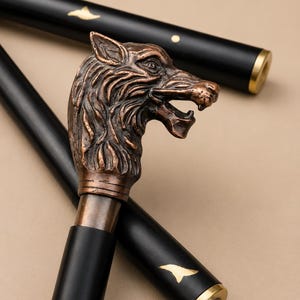 May include: A walking stick featuring a detailed bronze-coloured wolf head handle. The shaft is black with gold-coloured accents and a metallic tip. The design includes a sculpted wolf head with an open mouth and textured fur details on the handle.
