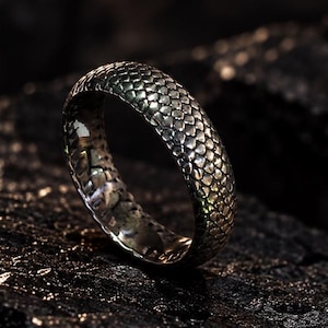 May include: A silver-toned ring featuring a textured, repeating scale pattern across its outer surface. The band has a rounded profile and a metallic finish that reflects light, showcasing a detailed, intricate design reminiscent of reptile skin.