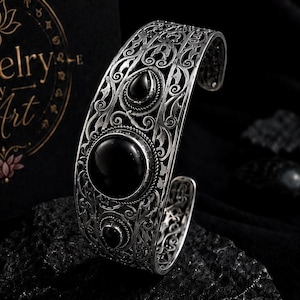 Puede incluir: Un brazalete de plata con intrincado trabajo de filigrana y tres piedras negras lisas y redondas dispuestas en fila vertical. Junto al brazalete hay una tarjeta negra con texto dorado que dice Jewelry By Art y un diseño de brújula.