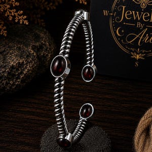 Puede incluir: Un brazalete de tono plateado con un diseño de banda de metal trenzado. El brazalete está acentuado con tres piedras lisas de color rojo oscuro en forma ovalada engastadas en el metal. La pieza se muestra vertical, exhibiendo su estructura abierta.