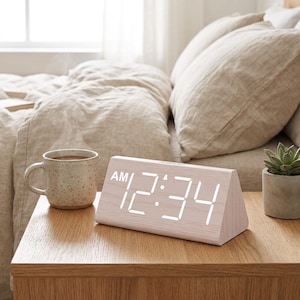 May include: A triangular digital alarm clock with a light wood grain finish and a bright white LED display showing the time 12:34 AM. The clock is positioned on a wooden surface next to a speckled ceramic mug and a small potted succulent plant.