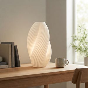 May include: A white, sculptural table lamp with a vertical, twisted, ribbed texture that emits a soft, warm glow. The lamp features an elongated, organic shape and is positioned next to a stack of books and a ceramic mug on a wooden surface.