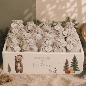 Op de afbeelding: Een witte houten kist gevuld met vierentwintig kleine beige stoffen zakjes met trekkoord, elk voorzien van een rond houten label genummerd van één tot vierentwintig. De kist is versierd met een beer met kerstmuts en de tekst ADVENTSKALENDER VON Niklas.