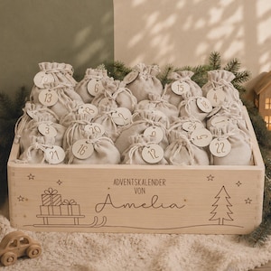 Puede incluir: Una caja de madera clara llena de veinticuatro pequeñas bolsas de tela beige con cordón, cada una con una etiqueta redonda de madera numerada del uno al veinticuatro. La caja está grabada con el texto ADVENTSKALENDER VON Amelia, un trineo con regalos y un pino.