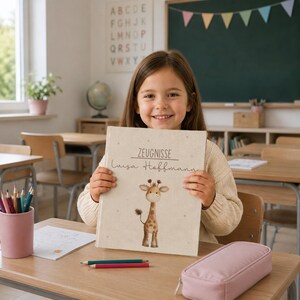 Puede incluir: Un libro de tapa dura beige con el texto Zeugnisse y Luisa Hoffmann impreso en la portada, que presenta una ilustración de una jirafa marrón y beige. La cubierta tiene una textura de tela clara con pequeños puntos grises.