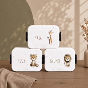 Könnte beinhalten: Drei weiße, rechteckige Brotdosen mit schwarzen Seitenclips. Jede Dose zeigt eine Tierillustration und einen Namen: eine Giraffe mit MAJA, ein Tiger mit LUCY und ein Löwe mit BRUNO. Der Markenname MEPAL ist auf jeder Dose aufgedruckt.