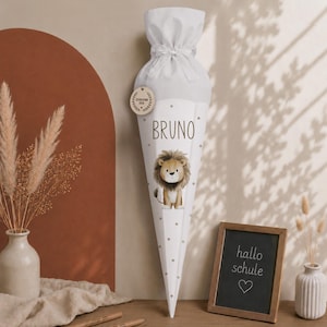 Op de afbeelding: Een witte, kegelvormige schoolcadeauzak met een bruine leeuwenillustratie, de naam BRUNO in zwarte tekst en een rond houten label met de tekst SCHULKIND 2026. De bovenkant is samengebonden met een wit lint en heeft lichtgrijze stippen.