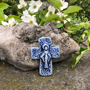 May include: A blue ceramic cross with a white figure of the Virgin Mary in the center. The cross is decorated with a floral design and is surrounded by white dogwood blossoms.
