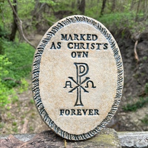 May include: A beige ceramic oval-shaped plaque with a black border and the text "MARKED AS CHRIST'S OWN" and "FOREVER" in black lettering. A black symbol with the letters "P", "A", "X", and "Ω" is in the center of the plaque.