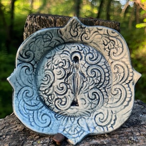 May include: A ceramic plate with a detailed design. The plate features a central figure surrounded by swirling patterns in shades of blue and white. The plate has a scalloped edge and is placed on a wooden surface.