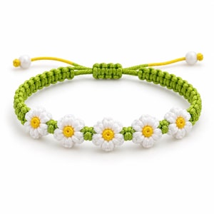 May include: A macrame bracelet featuring five white daisy flowers with yellow centres, woven from green cord. The adjustable band includes a sliding knot closure and two white beads at the ends of the yellow pull strings for a secure fit.