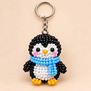 Könnte beinhalten: Ein Pinguin-Schlüsselanhänger aus kleinen schwarzen, weißen, blauen, gelben und rosafarbenen Perlen. Der Pinguin hat einen runden schwarzen Körper, einen weißen Bauch und ein weißes Gesicht, einen gelben Schnabel, schwarze Augen und eine blaue Schal.