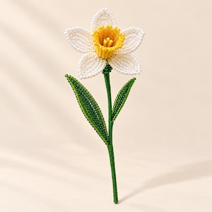 以下が含まれることがあります： 白い花びらと鮮やかな黄色の中心部を持つ、ビーズで作られた手作りの水仙の花。茎と2枚の尖った葉は小さな緑色のビーズで構成されており、春の花を詳細に表現した植物の造形物となっています。