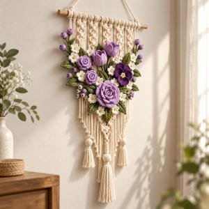 Könnte beinhalten: Ein Makramee-Wandbehang aus cremefarbener Baumwollkordel mit einem Holzstab und einem zentralen Arrangement aus gehäkelten Blumen in Lila-, Lavendel- und Weißtönen mit grünen Blättern, ergänzt durch dekorative Quasten und geknotete Details.