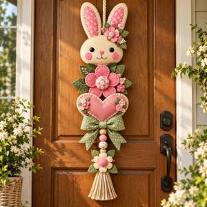 May include: A vertical hanging decoration featuring a felt rabbit head with pink floral ears, a large pink flower, a pink heart, a green polka-dot bow, wooden beads, and a bottom flower with a beige tassel, all suspended by a rope.
