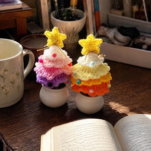 May include: Two crocheted Christmas tree ornaments in small white pots. The left tree features layers of white, pink, and purple yarn with small colorful pom-poms and a yellow star topper. The right tree has white, yellow, and orange layers with pom-poms and a yellow star.