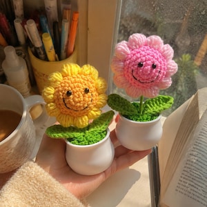 May include: Two small crocheted flowers in white pots. One flower is bright yellow and the other is light pink. Both flowers feature a round, textured face with black bead eyes and a stitched black smile. Each has green crocheted leaves.