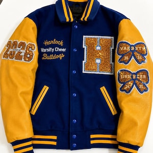 Könnte beinhalten: Eine blau-goldene Collegejacke mit gelben Lederärmeln, einem blauen Wollkörper und gestreiften Rippbündchen. Auf der Vorderseite befinden sich der Schriftzug Harlock Varsity Cheer Bulldogs, ein großer Buchstabe H, die Jahreszahl 2026 und zwei schleifenförmige Aufnäher.