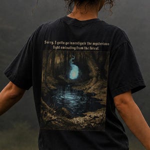 May include: A black short-sleeved t-shirt featuring a graphic print on the back of a dark forest scene with a glowing blue light above a pond. The text above the image reads, Sorry, I gotta go investigate the mysterious light emanating from the forest.