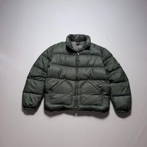 May include: A dark olive green puffer jacket featuring a high collar, a full-length front zipper, and two large front pockets. The jacket has a quilted design with horizontal baffles and elasticized cuffs, constructed from a textured fabric.