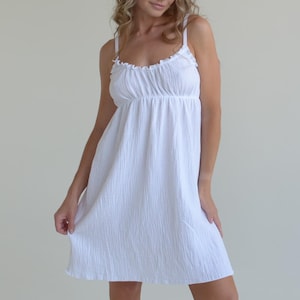 Pode incluir: Um vestido branco sem mangas com decote franzido com folhos e alças finas. A peça é feita de um tecido texturizado e leve que cai até à altura do joelho, criando uma silhueta simples e limpa.