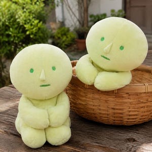 May include: Two pale lime green plush dolls with round heads, small pointed noses, and embroidered green eyes and mouths. One doll sits upright on a wooden surface, while the other rests inside a round woven wicker basket behind it.