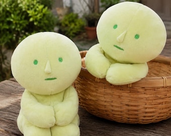 May include: Two pale lime green plush dolls with round heads, small pointed noses, and embroidered green eyes and mouths. One doll sits upright on a wooden surface, while the other rests inside a round woven wicker basket behind it.