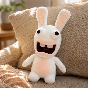 May include: A white plush rabbit toy with large, wide-set black eyes, two prominent front teeth, and a wide open mouth. The character features long ears with light pink inner sections and a matching light pink circular patch on its belly.