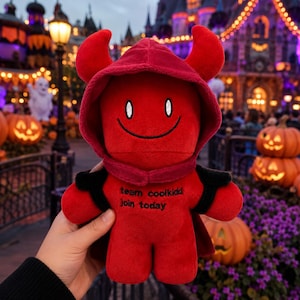 May include: A red plush toy featuring a hooded devil design with two horns, a stitched smiling face, and black embroidered text on the chest that reads team coolkidd join today. The soft fabric figure has a cape and rounded limbs.