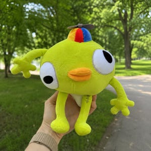May include: A plush toy featuring a bright lime green body with long, thin arms and legs. It has two large white eyes with black pupils, an orange beak, and a small propeller hat on top with red, blue, and yellow panels. A small stitch detail is visible.